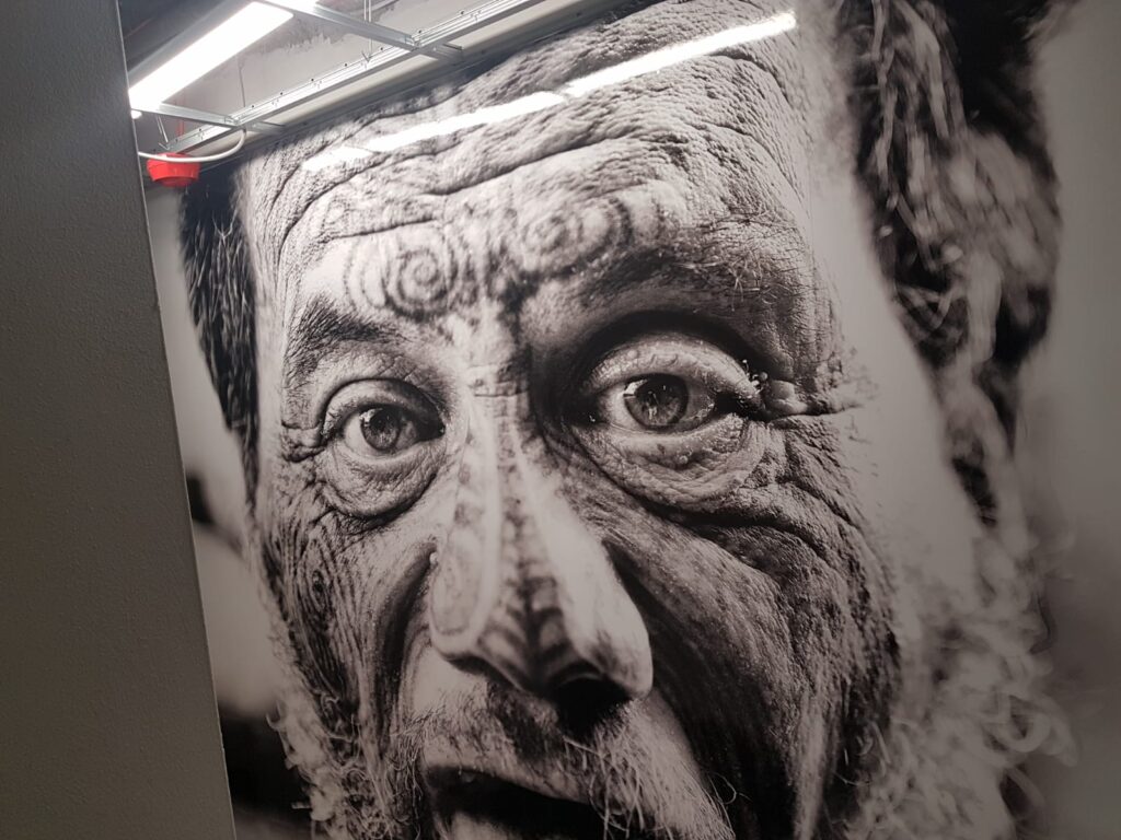 Großformatdruck in Flensburg 1 Großformatiges Schwarz-Weiß-Wandbild mit dem Nahaufnahme-Porträt eines älteren Maori-Mannes mit traditionellen Ta-Moko-Gesichtstätowierungen, an einer Innenraumwand unter Neonbeleuchtung angebracht.