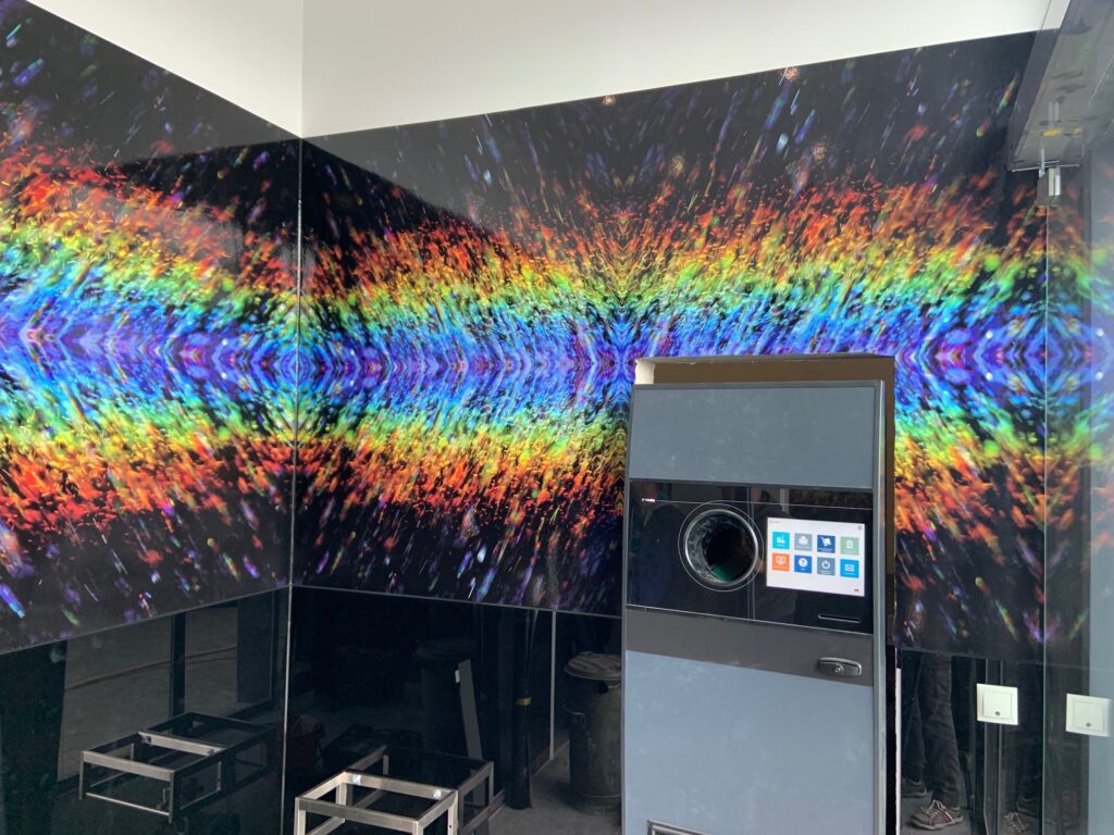 Großformatdruck in Kappeln 1 Raumecke mit großformatigem schwarzen Wandbild eines bunten Regenbogens aus abstrakten Lichtpartikeln, davor ein modernes Gerät mit Touchscreen-Display und metallischen Gestellen auf dem Boden.