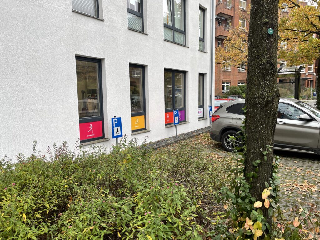 Firmenschilder im Kreis Rendsburg-Eckernförde 1 Außenfassade eines weißen Gebäudes in Flensburg mit bunten Piktogramm-Schildern zu den Themen Gesundheit und Mobilität sowie einem Behindertenparkplatz-Schild, davor ein bepflanzter Vorgarten und parkende Autos.