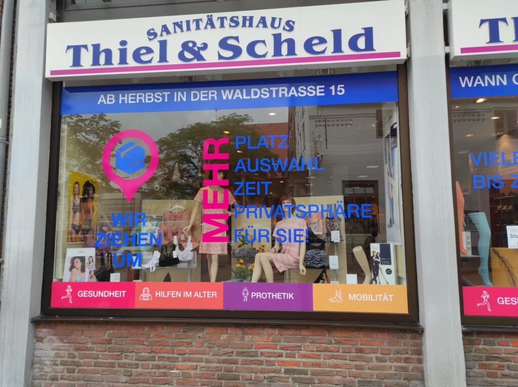 Großformatdruck in Süderbrarup 1 Schaufenster des Sanitätshauses Thiel & Scheld in Flensburg mit bunter Folienbeklebung, die einen Umzug in die Waldstraße 15 ankündigt und die Leistungsbereiche Gesundheit, Hilfen im Alter, Prothetik und Mobilität bewirbt.